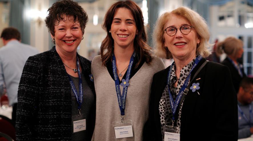Three women stand together with lanyards on, in a ballroom.