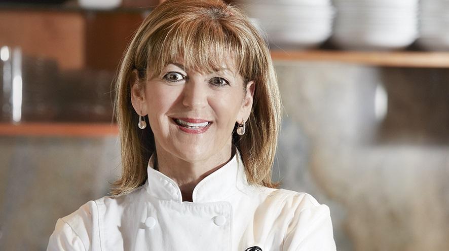 A woman with mid-length hair smiles. She's wearing a white chef's jacket.