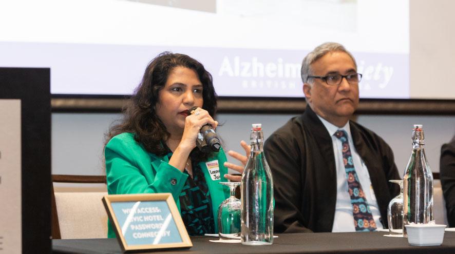 A South Asian woman and man sit at a table, speaking into microphones.