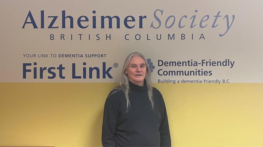 A man with long silver hair stands in front of a wall promoting services for people living with Alzheimer's disease and dementia.