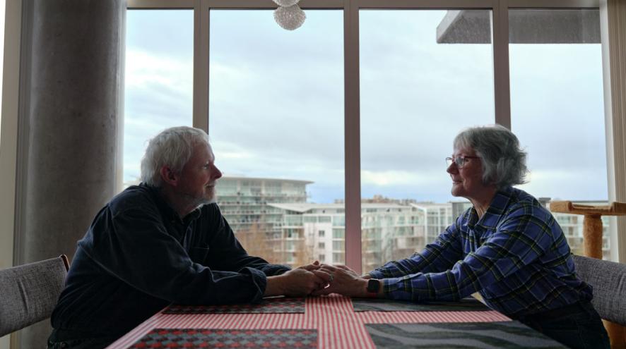 Two older adults sit across from each other at a table, holding hands in front of a large window overlooking the city.
