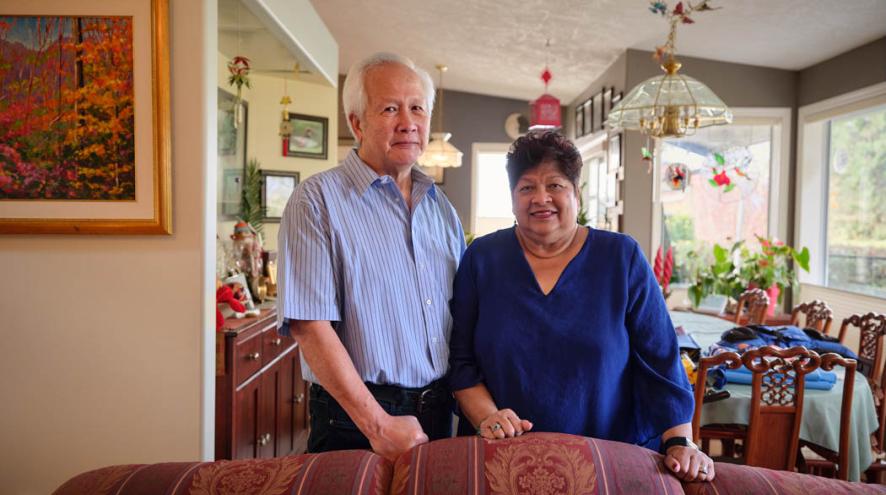 An older couple stand together behind a sofa in a warmly decorated home.
