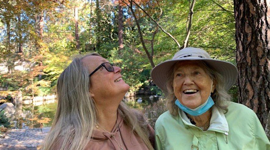 Two women stand in a forest on a sunny day, smiling. The woman on the left looks up. The woman on the right looks forward.