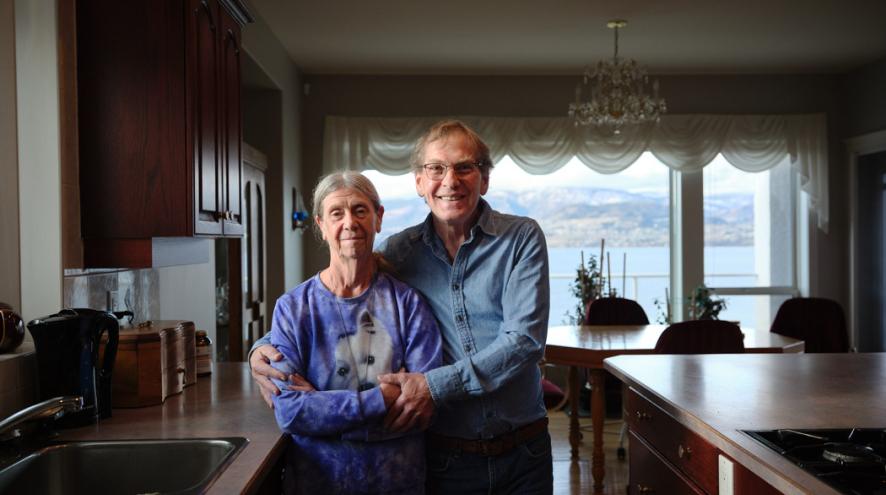 An older couple stand together in a bright kitchen, smiling at the camera.