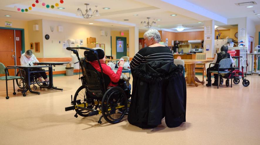 An older woman in a wheelchair sits with another older woman beside her in a spacious shared living area.