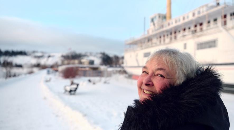 An older woman smiles outdoors in a snowy landscape, bundled in a winter coat with a large ship in the background.