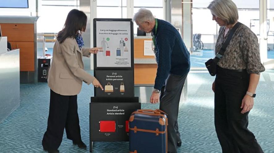 At the airport, an elder man is standing next to his travel companion. He is reaching for his luggage, while being assisted by an airport staff member to ensure his carryon luggage meets baggage allowance and standard size.