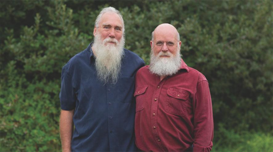 A male same sex couple stand together, looking at the camera with green foliage behind them