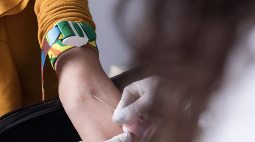 A health-care provider holds a needle against a person's inner arm below a tourniquet, in the moments before a blood draw. 