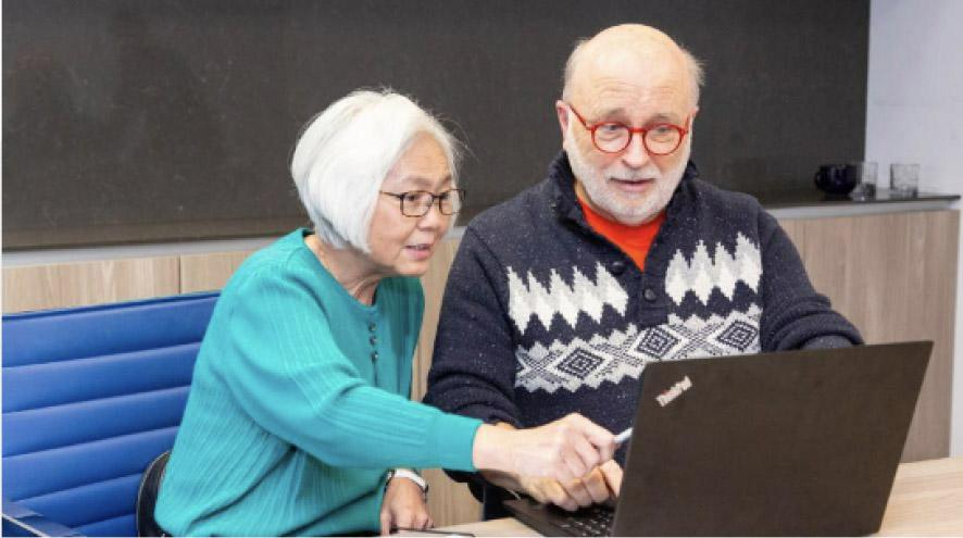 Two elderly people, wearing glasses, smile and point at their laptop screen.