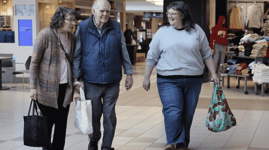 An elderly couple walk through a shopping mall, accompanied by a young woman. They carry shopping bags and are conversing. 