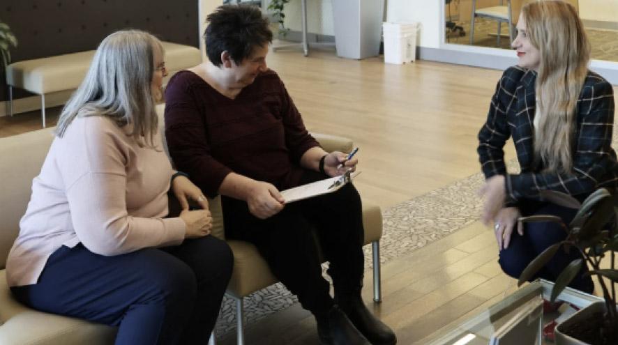 Two women sit in a bank lounge and speak with an employee. The employee crouches to be at eye level and makes eye contact in a friendly exchange.