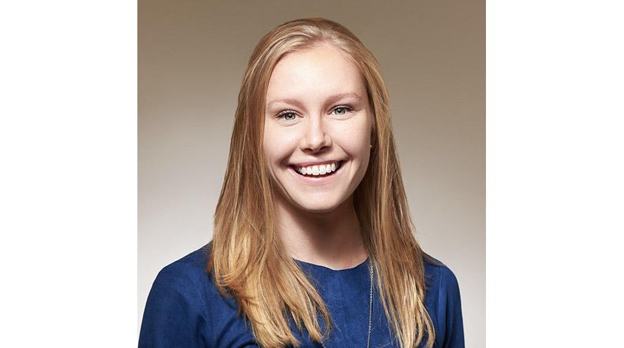 A young woman with copper hair smiles at the camera and poses for a headshot. She wears a blue sweater and stands against a plain background.