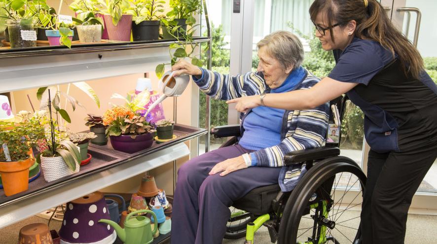 An older woman in a wheelchair receives assistance watering plants from a younger woman in scrubs.