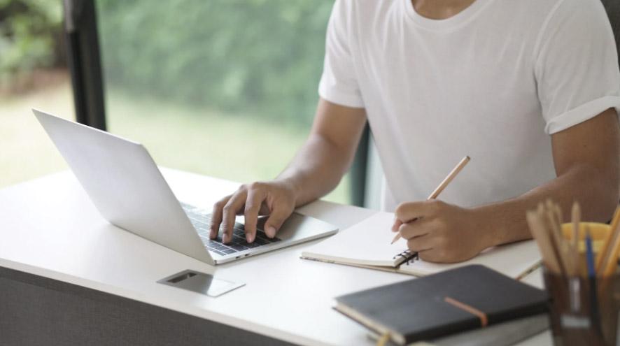 A man in a white shirt sits at a desk, working on his laptop while taking notes in a notebook.