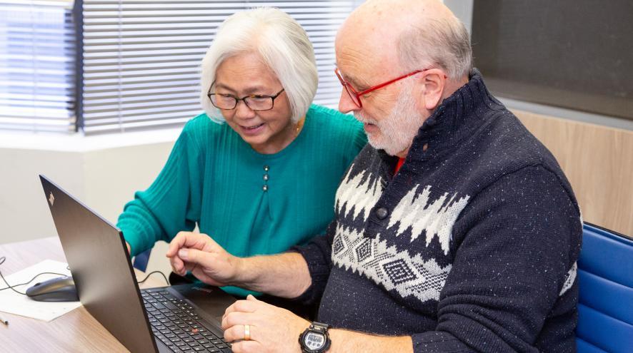 Two people sit close together in front of a laptop, engaged with what is occurring on screen. They smile with interest. One person is poised to type. 