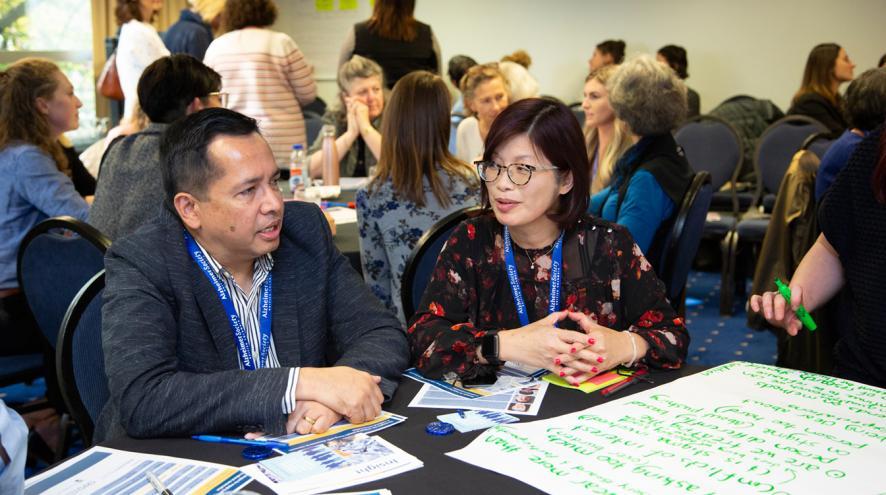 Two people sit in discussion at a round table, with notes in front of them and a round table of people in discussion behind them.