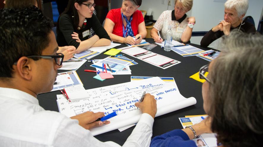 A person takes notes on oversized paper at a round table where a group of people are engaged in discussion in the background.