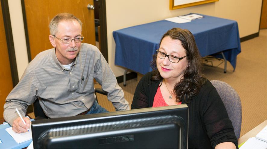 Two people are seated in front of a computer screen and engaged with what they're seeing. One person holds a pencil to paper, ready to take notes.