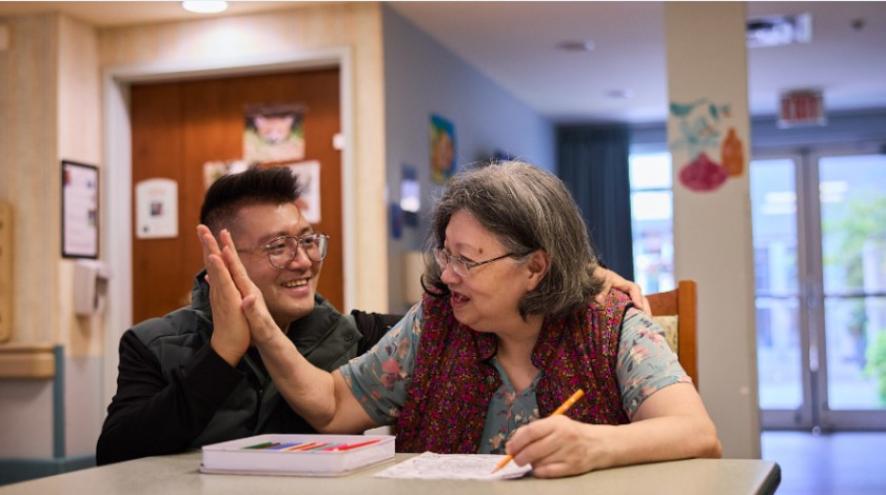A man and his mother sit at a desk smiling at each other. She writes with one hand and places her other palm to his.