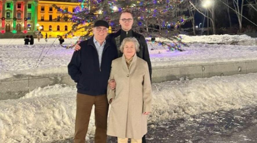 A man stands outside with his grandparents. It is snowing and they are dressed warmly and look happy.