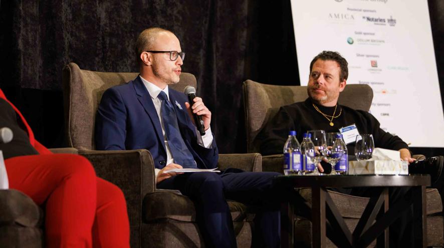 Panel discussion on stage: Two men converse in armchairs, one holding a microphone, with a woman nearby and sponsor backdrop behind them.