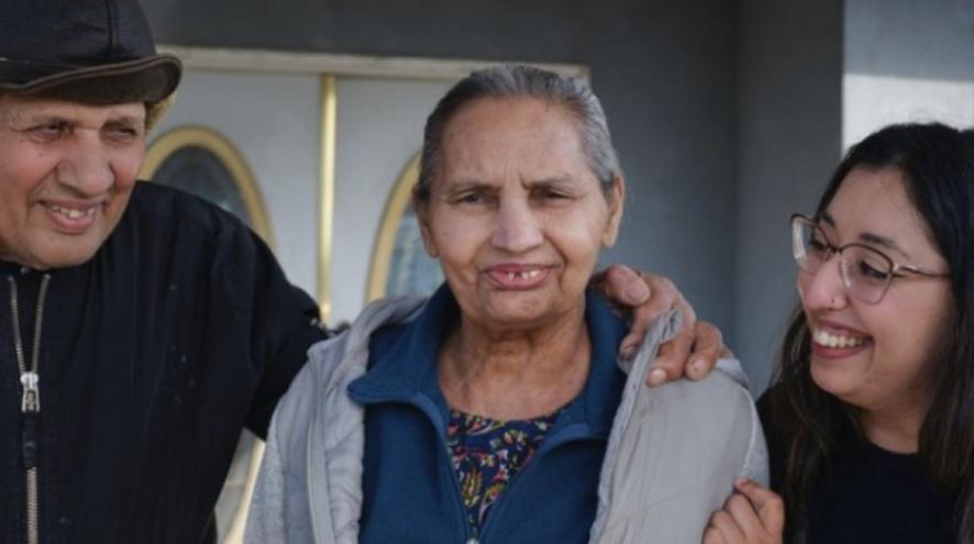 A man, a woman and a young woman stand together. The young woman is looking at the older woman and smiling.