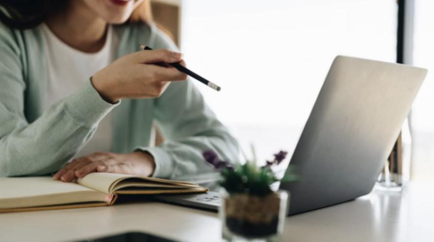 a close up of a woman wearing a green cardigan pointing a marker towards a laptop. 