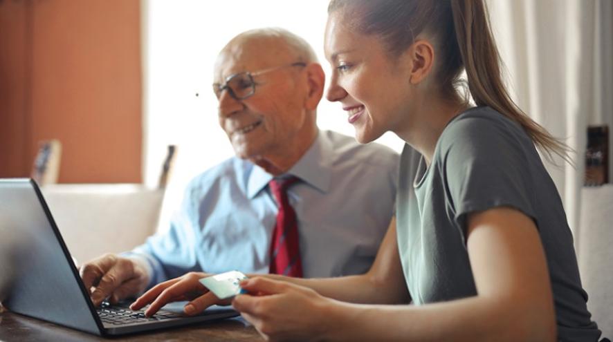 A younger woman sits at a laptop with an elderly man. They are both looking at the screen and smiling.