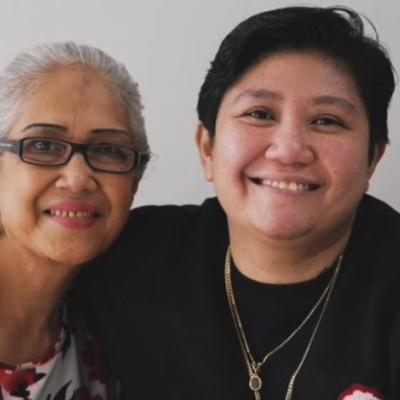 An older woman with gray hair and glasses, smiles and poses close to a younger person with short dark hair