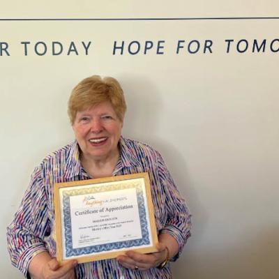 Happy older woman stands in front of a white office wall holding a certificate of recognition 