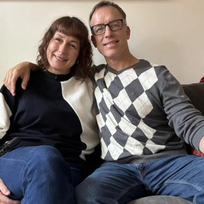 Karina and Andrew Reid are sitting next to each other on their living room couch, with Andrew's arm on Karina's shoulder.