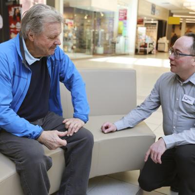 Inside a shopping mall, a gentleman is sitting on a bench while talking to a worker. The worker is kneeling, speaking to the man at eye level.