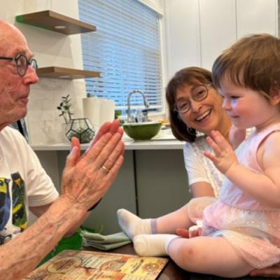 Lived experience Dan is playfully clapping his hands in front of his grand draughter, who is sitting on the dining table and held by her grandmother.