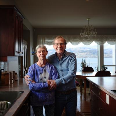 An older couple stand together in a bright kitchen, smiling at the camera.