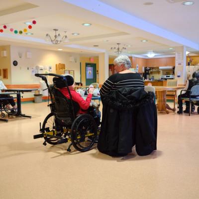 An older woman in a wheelchair sits with another older woman beside her in a spacious shared living area.