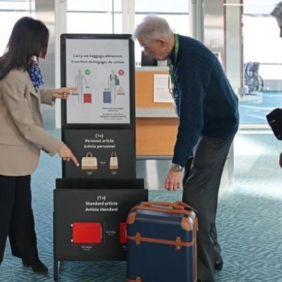 At the airport, an elder man is standing next to his travel companion. He is reaching for his luggage, while being assisted by an airport staff member to ensure his carryon luggage meets baggage allowance and standard size.
