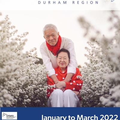 Man with red scarf wearing a white shirt with arms around the shoulders of a woman with a red shirt and white pants. The man is standing and the woman is seated surrounded by snow covered trees.