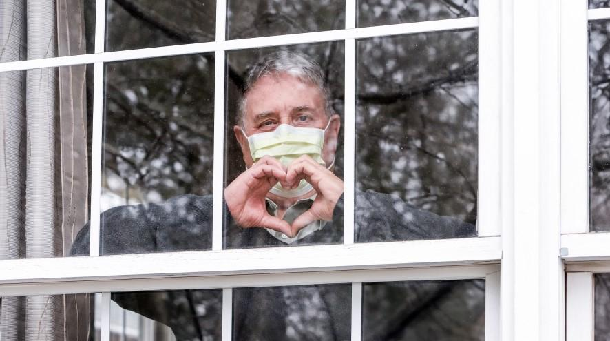 Senior man wearing a mask showing the heart symbol from inside his room