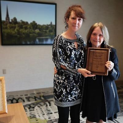 Photo of Riba being presented with the Pauline Spatz Leadership Award, a wooden plaque, by ASNB staff