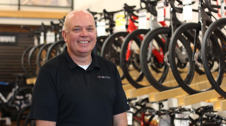 A bald man in a black polo tshirt smiling in front of a row of bicycles on display. His shirt logo reads "Cyclesmith"