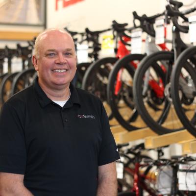 A bald man in a black polo tshirt smiling in front of a row of bicycles on display. His shirt logo reads "Cyclesmith"