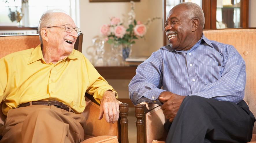 Elderly couple relaxing side by side in armchairs. The man on the left is light skinned and wearing a yellow shirt, and on the right is a Black man with dark brown skin wearing a pale blue shirt.