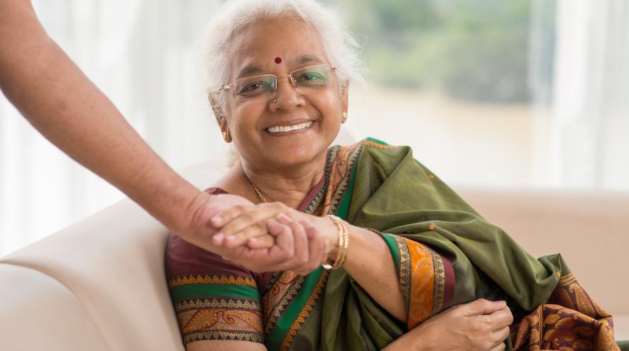 Elderly Indian woman wearing a bindi and a green saree, smiling as her hand is clasped by a person standing out of frame.