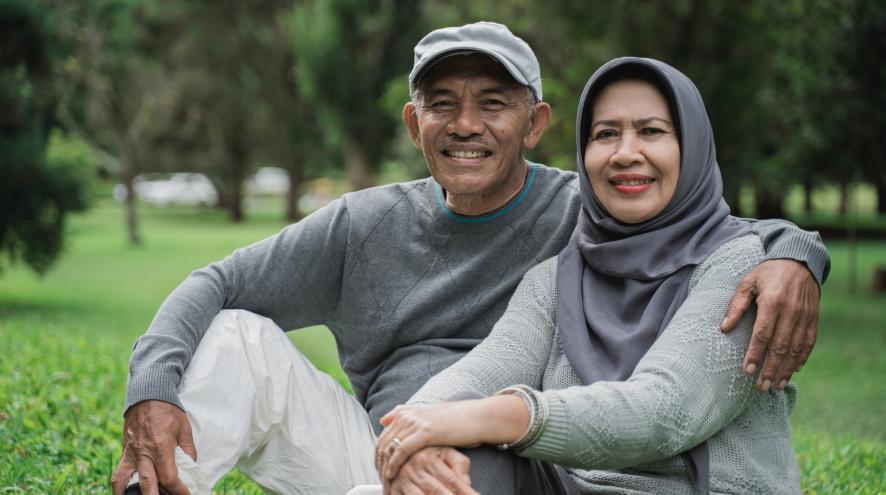 Elderly man and woman sitting together. The woman has light skin and wears a grey hijab. The man has darker brown skin and is wearing a baseball cap.