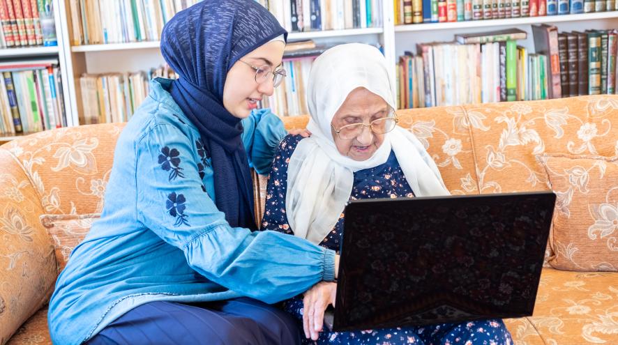 Young Muslim woman wearing a dark blue hijab over a lighter blue dress, helping an elderly woman in a white head scarf with a laptop.