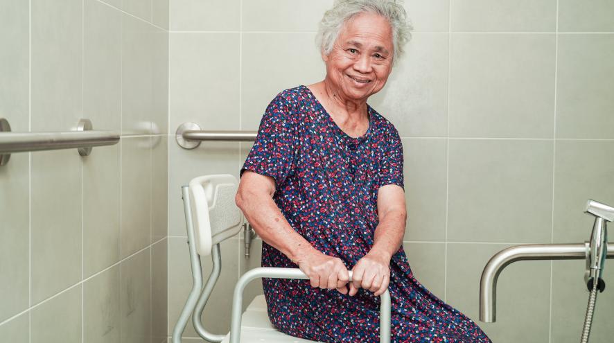 Elderly Asian woman in a patterned blue dress, seated on an assistive shower chair.