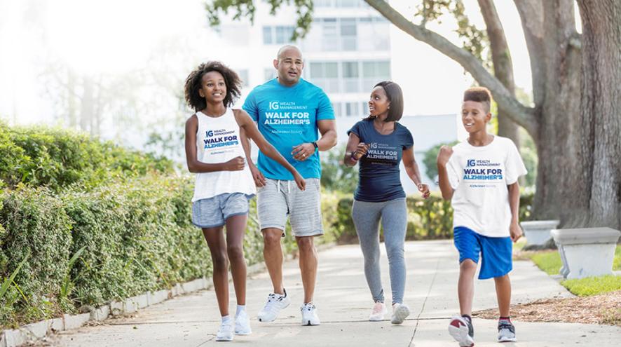 Four people - a tall Black man with a bald head, a Black woman with straight dark hair, a Black girl with curly hair, an a younger Black boy with short cropped curly hair - all dressed in IGWM Walk for Alzheimer's shirts, walking outdoors on a downtown sidewalk, surrounded by shrubbery and trees. High-rise buildings can be seen in the background.