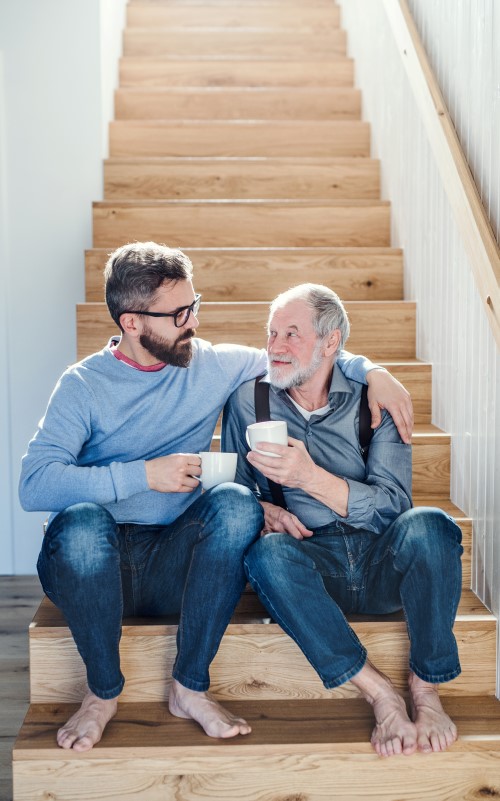 A young man and a senior man sitting on the stairs together and having coffee, with the young man putting his arm around the senior man's shoulders.
