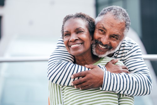 Senior man hugging his wife from behind, who is sitting down and also smiling.